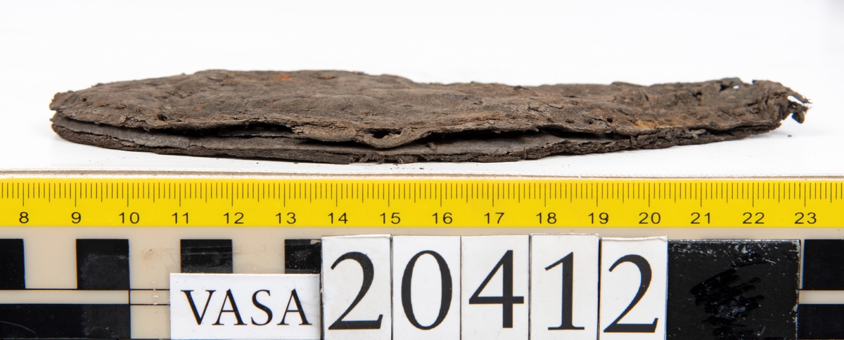 Dokumentation 1997:
Delar och fragment från sko: sulor, slejf, klack, del av holtrand mm. Totalt 23 stycken delar och fragment.
Del av klack bestående av fyra klacklappar. Klacken har kvarsittande skopligg och tråd. Lädret har rostfärgade utfällningar.
Främre partiet av två sulor, sulornas hälparti saknas. Sulorna hålls samman med kvarsittande tråd. Lädret är spaltat.
En fragmentarisk och spaltad sula. Sulan har kvarsittande tråd. Lädret har rostfärgade utfällningar.
En slejf. I slejfens ena ände finns ett snörhål. Runt kanterna löper hål efter stygn.
En textilfiber.

Dokumentation 2022:
Slejf som tillhör bakkappan av sko med fyndnummer 20424.
För mer information, se Uppritning under Länkade filer.

Text in english:
This is the pieced latchet belonging to the shoe quarter 20424.
For more information, go to Länkade filer: Uppritning