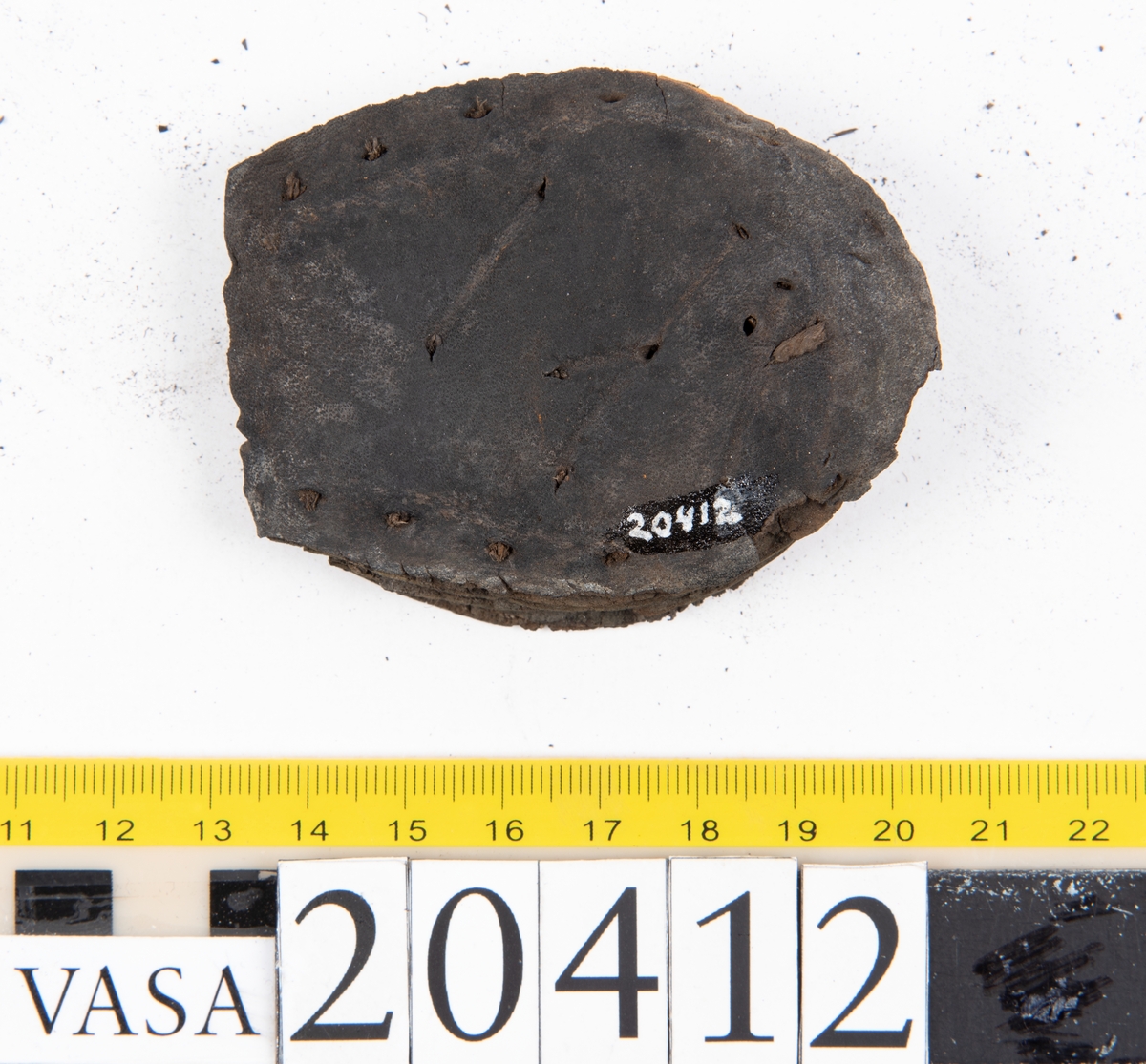 Dokumentation 1997:
Delar och fragment från sko: sulor, slejf, klack, del av holtrand mm. Totalt 23 stycken delar och fragment.
Del av klack bestående av fyra klacklappar. Klacken har kvarsittande skopligg och tråd. Lädret har rostfärgade utfällningar.
Främre partiet av två sulor, sulornas hälparti saknas. Sulorna hålls samman med kvarsittande tråd. Lädret är spaltat.
En fragmentarisk och spaltad sula. Sulan har kvarsittande tråd. Lädret har rostfärgade utfällningar.
En slejf. I slejfens ena ände finns ett snörhål. Runt kanterna löper hål efter stygn.
En textilfiber.

Dokumentation 2022:
Slejf som tillhör bakkappan av sko med fyndnummer 20424.
För mer information, se Uppritning under Länkade filer.

Text in english:
This is the pieced latchet belonging to the shoe quarter 20424.
For more information, go to Länkade filer: Uppritning