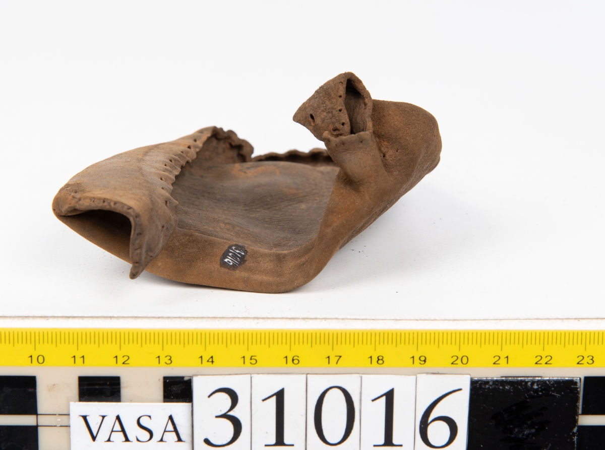 Ovanläder och bakkappa till sko. Längs med ovanlädrets ytterkant löper en enkel rad med stygnshål. Narvytan är välbevarad och har en rutmönstrad struktur. Bakkappans nedre kant är vikt. Kanten har legat invikt och suttit fästad i sulpaketet.