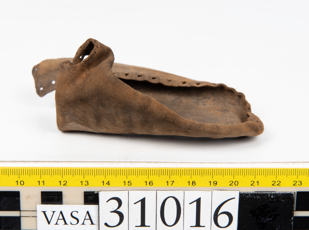 Ovanläder och bakkappa till sko. Längs med ovanlädrets ytterkant löper en enkel rad med stygnshål. Narvytan är välbevarad och har en rutmönstrad struktur. Bakkappans nedre kant är vikt. Kanten har legat invikt och suttit fästad i sulpaketet.