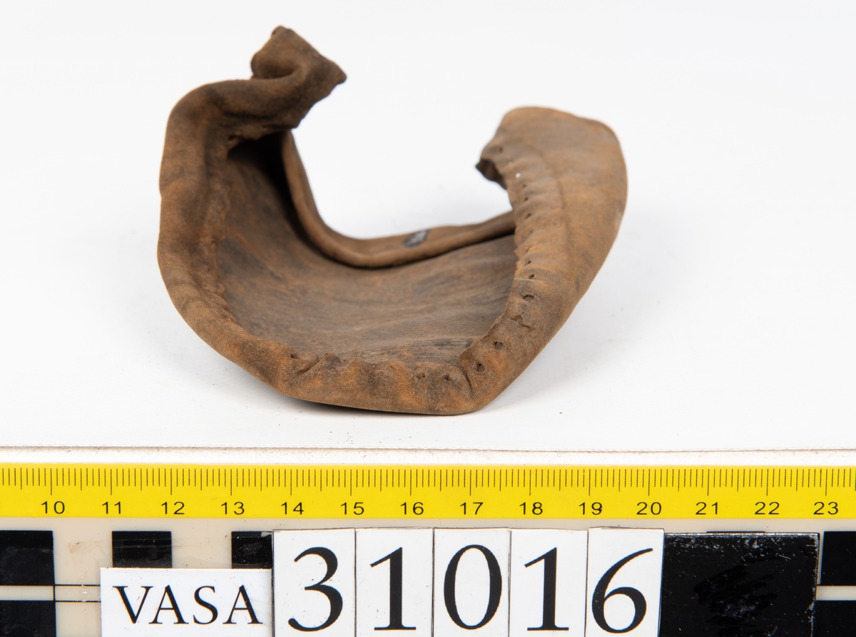 Ovanläder och bakkappa till sko. Längs med ovanlädrets ytterkant löper en enkel rad med stygnshål. Narvytan är välbevarad och har en rutmönstrad struktur. Bakkappans nedre kant är vikt. Kanten har legat invikt och suttit fästad i sulpaketet.