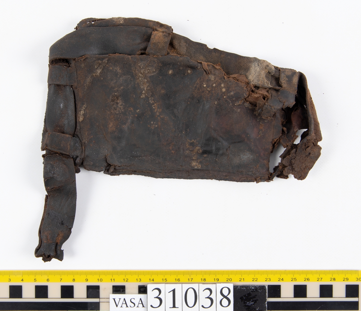 Delar av en väska. Totalt rör det sig om fyra delar, varav två lösa delar av en läderrem.
Halvcirkelformat skuret läderstycke, fyndnummer 31038. Troligtvis utgör läderstycket en del av bottnen och sidostycket till en väska.  Längs med ytterkanten syns ritsade skåror som är ritsade två och två. Genom skårorna löper en, ca 20 mm bred, läderrem. Remmen har troligtvis fungerat som axelrem, eventuellt livrem. Ena änden av remmen är dubbelvikt. Där remmen vikts finns ett runt hål. Kring hålet finns intryck och rostliknande utfällningar, troligtvis efter ett spänne. På motsatta sidan syns hål efter stygn som löper i formen av en rektangel. På läderstyckets nedre del finns runda, hårda, grönfärgade blåsor.
Delar av läderstycket är fragmentariskt eller saknas och lädret har rostfärgade utfällningar.
Sidostycke eller del av lock till en väska, fyndnummer 31038:1. Lädret är skuret i ett rektangulärt stycke. Runt ytterkanterna löper stygnshål. I ett av hörnen finns en rektangulär skåra. På läderstycket syns svaga intryck i form av cirklar.
Del av rem, fyndnummer 31038:2. I ena änden finns stygnshål som löper i formen av en rektangel.
Del av rem, fyndnummer 31038:3. Remmen dubbelvikt och tycks sakna stygnshål.
Lädret är på det hela taget kraftigt och välbevarat.