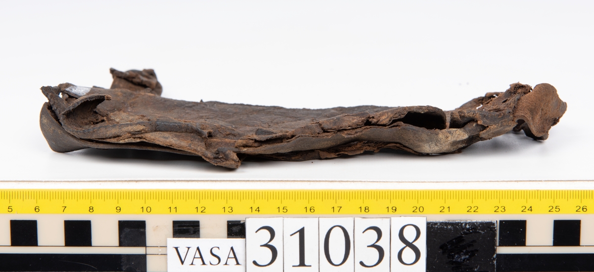Delar av en väska. Totalt rör det sig om fyra delar, varav två lösa delar av en läderrem.
Halvcirkelformat skuret läderstycke, fyndnummer 31038. Troligtvis utgör läderstycket en del av bottnen och sidostycket till en väska.  Längs med ytterkanten syns ritsade skåror som är ritsade två och två. Genom skårorna löper en, ca 20 mm bred, läderrem. Remmen har troligtvis fungerat som axelrem, eventuellt livrem. Ena änden av remmen är dubbelvikt. Där remmen vikts finns ett runt hål. Kring hålet finns intryck och rostliknande utfällningar, troligtvis efter ett spänne. På motsatta sidan syns hål efter stygn som löper i formen av en rektangel. På läderstyckets nedre del finns runda, hårda, grönfärgade blåsor.
Delar av läderstycket är fragmentariskt eller saknas och lädret har rostfärgade utfällningar.
Sidostycke eller del av lock till en väska, fyndnummer 31038:1. Lädret är skuret i ett rektangulärt stycke. Runt ytterkanterna löper stygnshål. I ett av hörnen finns en rektangulär skåra. På läderstycket syns svaga intryck i form av cirklar.
Del av rem, fyndnummer 31038:2. I ena änden finns stygnshål som löper i formen av en rektangel.
Del av rem, fyndnummer 31038:3. Remmen dubbelvikt och tycks sakna stygnshål.
Lädret är på det hela taget kraftigt och välbevarat.