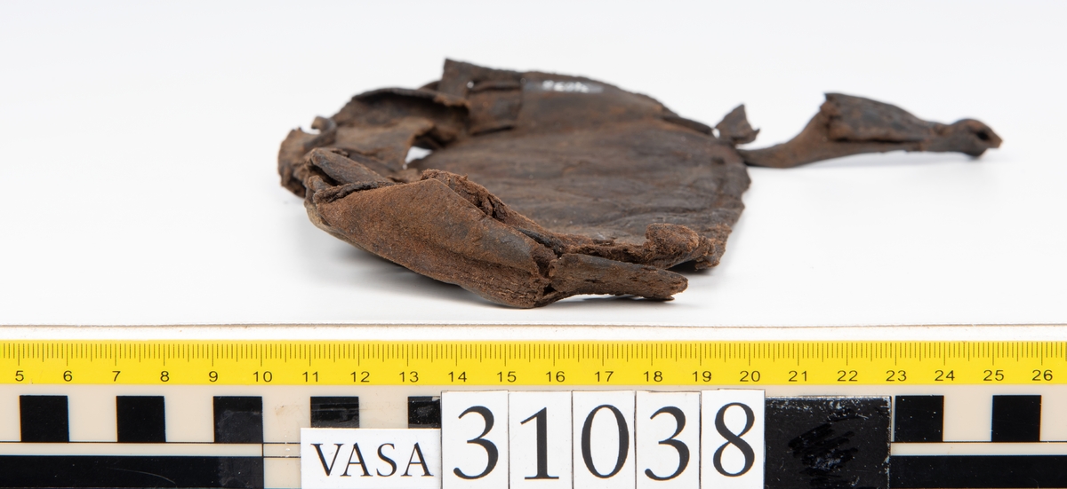 Delar av en väska. Totalt rör det sig om fyra delar, varav två lösa delar av en läderrem.
Halvcirkelformat skuret läderstycke, fyndnummer 31038. Troligtvis utgör läderstycket en del av bottnen och sidostycket till en väska.  Längs med ytterkanten syns ritsade skåror som är ritsade två och två. Genom skårorna löper en, ca 20 mm bred, läderrem. Remmen har troligtvis fungerat som axelrem, eventuellt livrem. Ena änden av remmen är dubbelvikt. Där remmen vikts finns ett runt hål. Kring hålet finns intryck och rostliknande utfällningar, troligtvis efter ett spänne. På motsatta sidan syns hål efter stygn som löper i formen av en rektangel. På läderstyckets nedre del finns runda, hårda, grönfärgade blåsor.
Delar av läderstycket är fragmentariskt eller saknas och lädret har rostfärgade utfällningar.
Sidostycke eller del av lock till en väska, fyndnummer 31038:1. Lädret är skuret i ett rektangulärt stycke. Runt ytterkanterna löper stygnshål. I ett av hörnen finns en rektangulär skåra. På läderstycket syns svaga intryck i form av cirklar.
Del av rem, fyndnummer 31038:2. I ena änden finns stygnshål som löper i formen av en rektangel.
Del av rem, fyndnummer 31038:3. Remmen dubbelvikt och tycks sakna stygnshål.
Lädret är på det hela taget kraftigt och välbevarat.