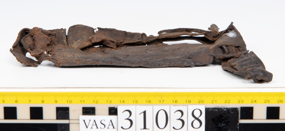 Delar av en väska. Totalt rör det sig om fyra delar, varav två lösa delar av en läderrem.
Halvcirkelformat skuret läderstycke, fyndnummer 31038. Troligtvis utgör läderstycket en del av bottnen och sidostycket till en väska.  Längs med ytterkanten syns ritsade skåror som är ritsade två och två. Genom skårorna löper en, ca 20 mm bred, läderrem. Remmen har troligtvis fungerat som axelrem, eventuellt livrem. Ena änden av remmen är dubbelvikt. Där remmen vikts finns ett runt hål. Kring hålet finns intryck och rostliknande utfällningar, troligtvis efter ett spänne. På motsatta sidan syns hål efter stygn som löper i formen av en rektangel. På läderstyckets nedre del finns runda, hårda, grönfärgade blåsor.
Delar av läderstycket är fragmentariskt eller saknas och lädret har rostfärgade utfällningar.
Sidostycke eller del av lock till en väska, fyndnummer 31038:1. Lädret är skuret i ett rektangulärt stycke. Runt ytterkanterna löper stygnshål. I ett av hörnen finns en rektangulär skåra. På läderstycket syns svaga intryck i form av cirklar.
Del av rem, fyndnummer 31038:2. I ena änden finns stygnshål som löper i formen av en rektangel.
Del av rem, fyndnummer 31038:3. Remmen dubbelvikt och tycks sakna stygnshål.
Lädret är på det hela taget kraftigt och välbevarat.