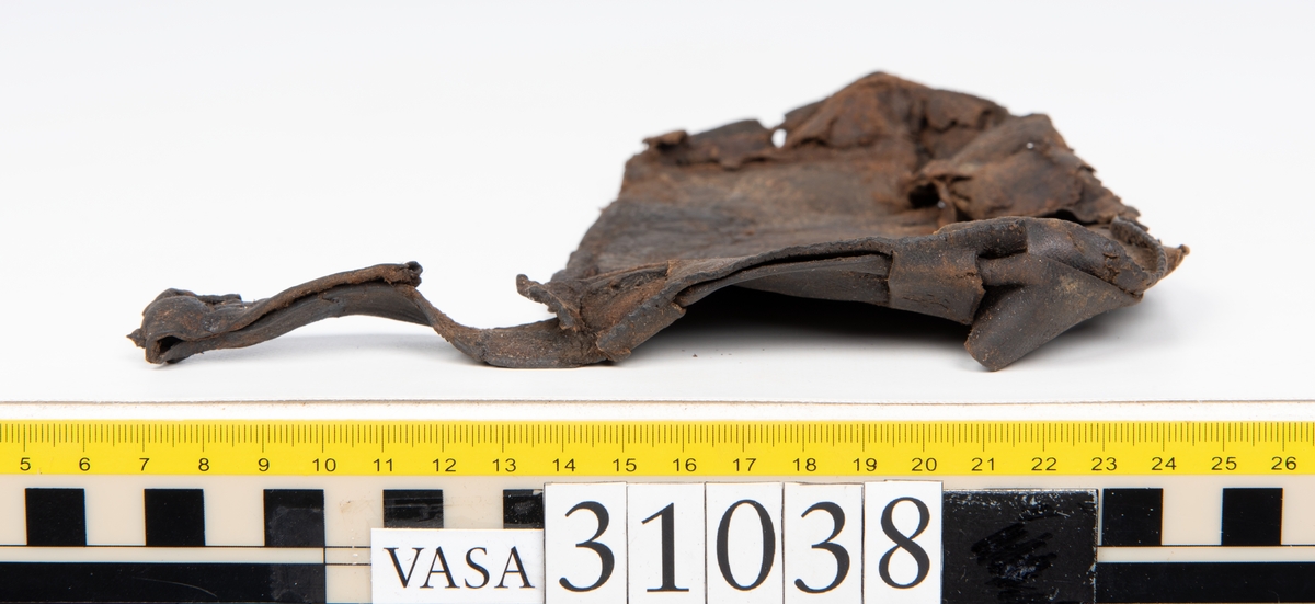 Delar av en väska. Totalt rör det sig om fyra delar, varav två lösa delar av en läderrem.
Halvcirkelformat skuret läderstycke, fyndnummer 31038. Troligtvis utgör läderstycket en del av bottnen och sidostycket till en väska.  Längs med ytterkanten syns ritsade skåror som är ritsade två och två. Genom skårorna löper en, ca 20 mm bred, läderrem. Remmen har troligtvis fungerat som axelrem, eventuellt livrem. Ena änden av remmen är dubbelvikt. Där remmen vikts finns ett runt hål. Kring hålet finns intryck och rostliknande utfällningar, troligtvis efter ett spänne. På motsatta sidan syns hål efter stygn som löper i formen av en rektangel. På läderstyckets nedre del finns runda, hårda, grönfärgade blåsor.
Delar av läderstycket är fragmentariskt eller saknas och lädret har rostfärgade utfällningar.
Sidostycke eller del av lock till en väska, fyndnummer 31038:1. Lädret är skuret i ett rektangulärt stycke. Runt ytterkanterna löper stygnshål. I ett av hörnen finns en rektangulär skåra. På läderstycket syns svaga intryck i form av cirklar.
Del av rem, fyndnummer 31038:2. I ena änden finns stygnshål som löper i formen av en rektangel.
Del av rem, fyndnummer 31038:3. Remmen dubbelvikt och tycks sakna stygnshål.
Lädret är på det hela taget kraftigt och välbevarat.