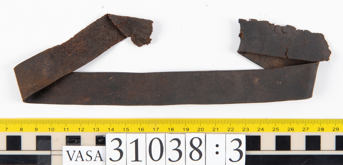 Delar av en väska. Totalt rör det sig om fyra delar, varav två lösa delar av en läderrem.
Halvcirkelformat skuret läderstycke, fyndnummer 31038. Troligtvis utgör läderstycket en del av bottnen och sidostycket till en väska.  Längs med ytterkanten syns ritsade skåror som är ritsade två och två. Genom skårorna löper en, ca 20 mm bred, läderrem. Remmen har troligtvis fungerat som axelrem, eventuellt livrem. Ena änden av remmen är dubbelvikt. Där remmen vikts finns ett runt hål. Kring hålet finns intryck och rostliknande utfällningar, troligtvis efter ett spänne. På motsatta sidan syns hål efter stygn som löper i formen av en rektangel. På läderstyckets nedre del finns runda, hårda, grönfärgade blåsor.
Delar av läderstycket är fragmentariskt eller saknas och lädret har rostfärgade utfällningar.
Sidostycke eller del av lock till en väska, fyndnummer 31038:1. Lädret är skuret i ett rektangulärt stycke. Runt ytterkanterna löper stygnshål. I ett av hörnen finns en rektangulär skåra. På läderstycket syns svaga intryck i form av cirklar.
Del av rem, fyndnummer 31038:2. I ena änden finns stygnshål som löper i formen av en rektangel.
Del av rem, fyndnummer 31038:3. Remmen dubbelvikt och tycks sakna stygnshål.
Lädret är på det hela taget kraftigt och välbevarat.