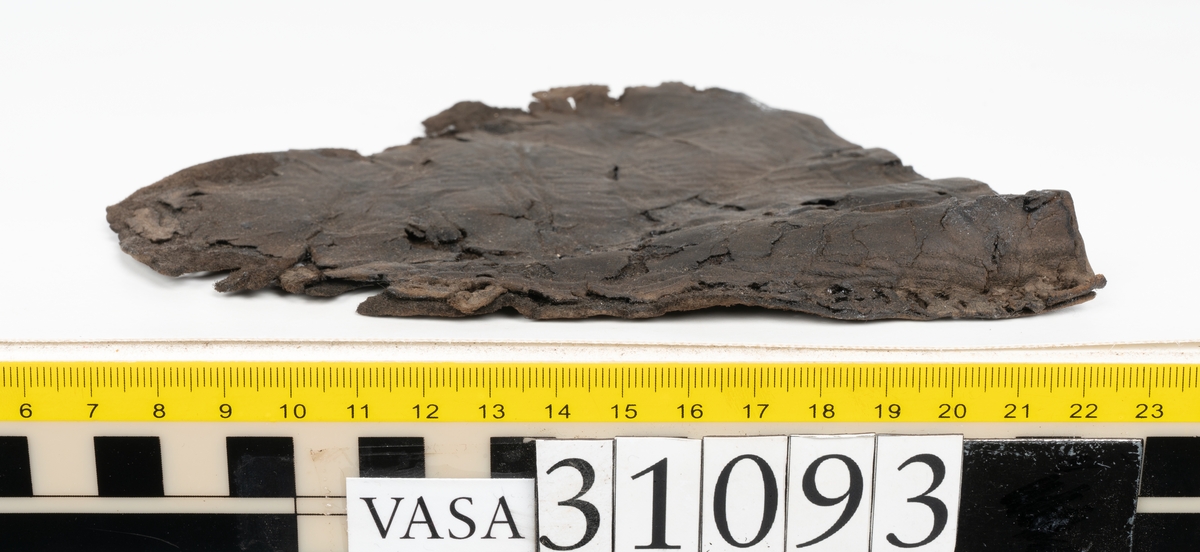 Ett ovanläder till en sko. Ovanlädrets övre kant är rakt avskuren och saknar tunga. Nedanför den övre kanten sitter två snörhål. Längs med ytterkanterna löper stygnshål. Ena ytterkanten är fragmentarisk.