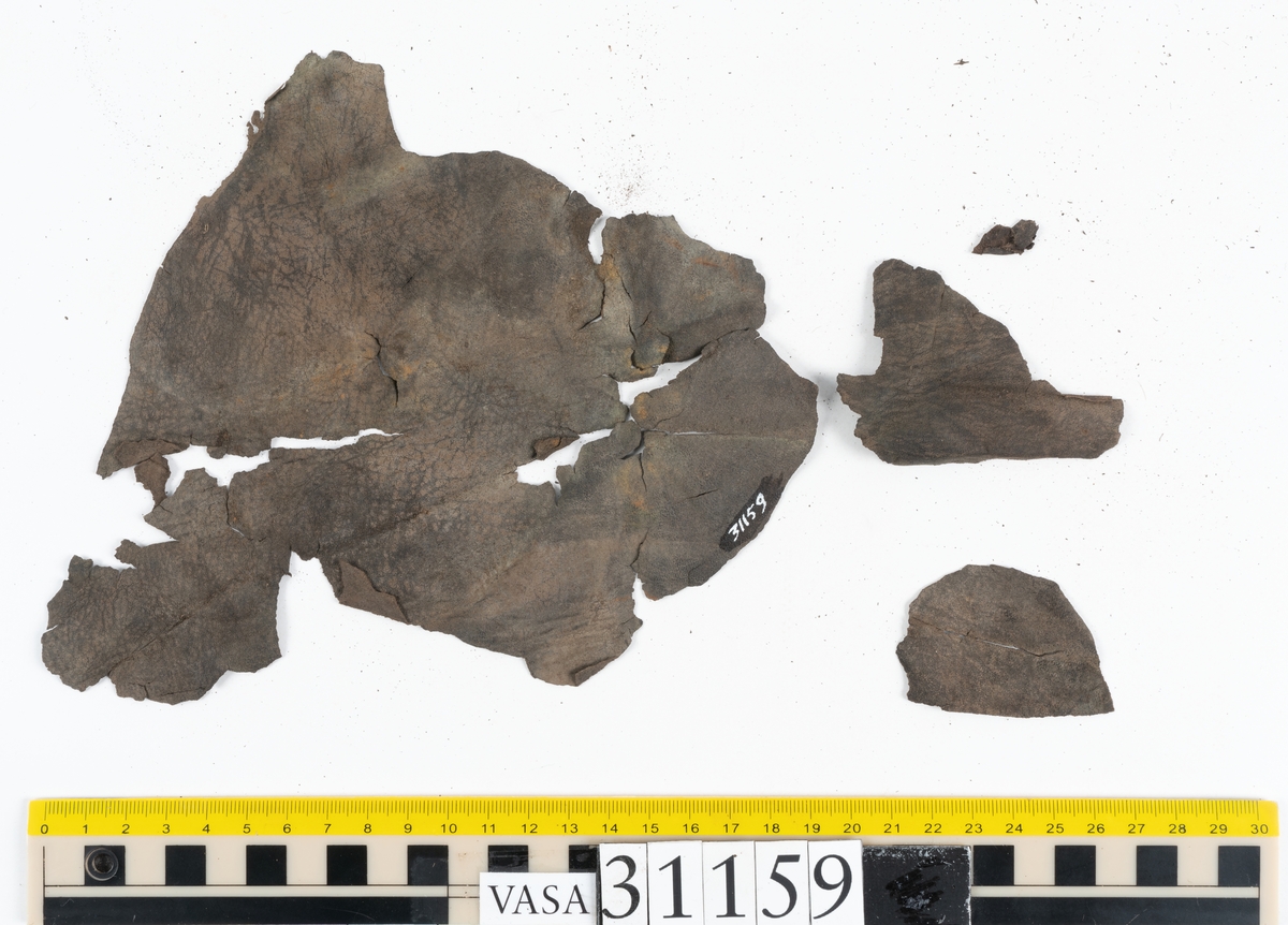 Två större läderstycken samt ett tjugotal mindre fragment, troligtvis från stövelskaft. Lädret har rostliknande utfällningar samt är spaltat och sprött.