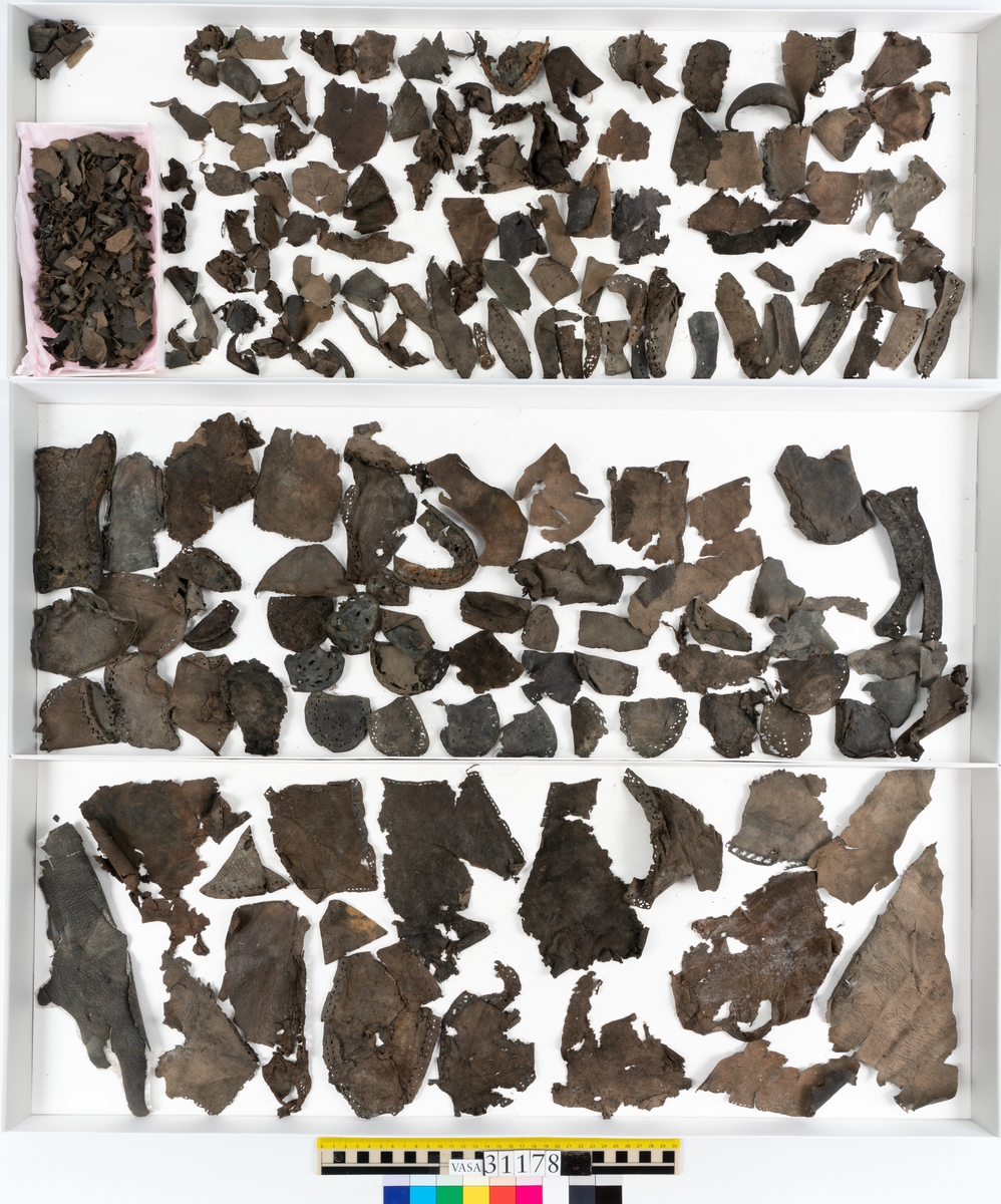 Runt 400 delar och fragment från skor i form av sulor, inlägg, ovanläder, klack, sidostycken, bakkappor, träpliggar, tråd mm. Troligtvis rör det sig om delar och fragment från flera skor och/eller stövlar. Delar av lädret har förstärkts med lim av konserveringsenheten. Merparten av lädret är spaltat samt har hål efter träpligg eller stygn.