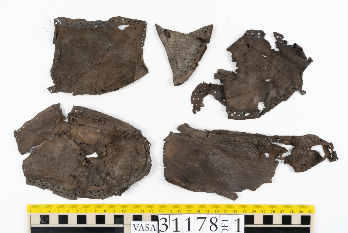 Runt 400 delar och fragment från skor i form av sulor, inlägg, ovanläder, klack, sidostycken, bakkappor, träpliggar, tråd mm. Troligtvis rör det sig om delar och fragment från flera skor och/eller stövlar. Delar av lädret har förstärkts med lim av konserveringsenheten. Merparten av lädret är spaltat samt har hål efter träpligg eller stygn.