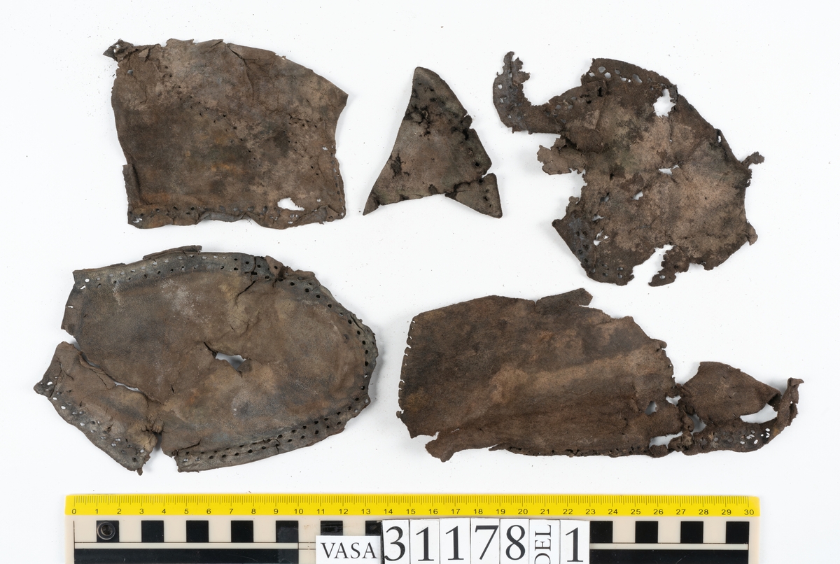 Runt 400 delar och fragment från skor i form av sulor, inlägg, ovanläder, klack, sidostycken, bakkappor, träpliggar, tråd mm. Troligtvis rör det sig om delar och fragment från flera skor och/eller stövlar. Delar av lädret har förstärkts med lim av konserveringsenheten. Merparten av lädret är spaltat samt har hål efter träpligg eller stygn.