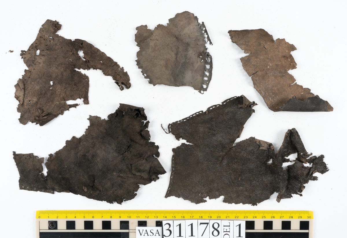 Runt 400 delar och fragment från skor i form av sulor, inlägg, ovanläder, klack, sidostycken, bakkappor, träpliggar, tråd mm. Troligtvis rör det sig om delar och fragment från flera skor och/eller stövlar. Delar av lädret har förstärkts med lim av konserveringsenheten. Merparten av lädret är spaltat samt har hål efter träpligg eller stygn.