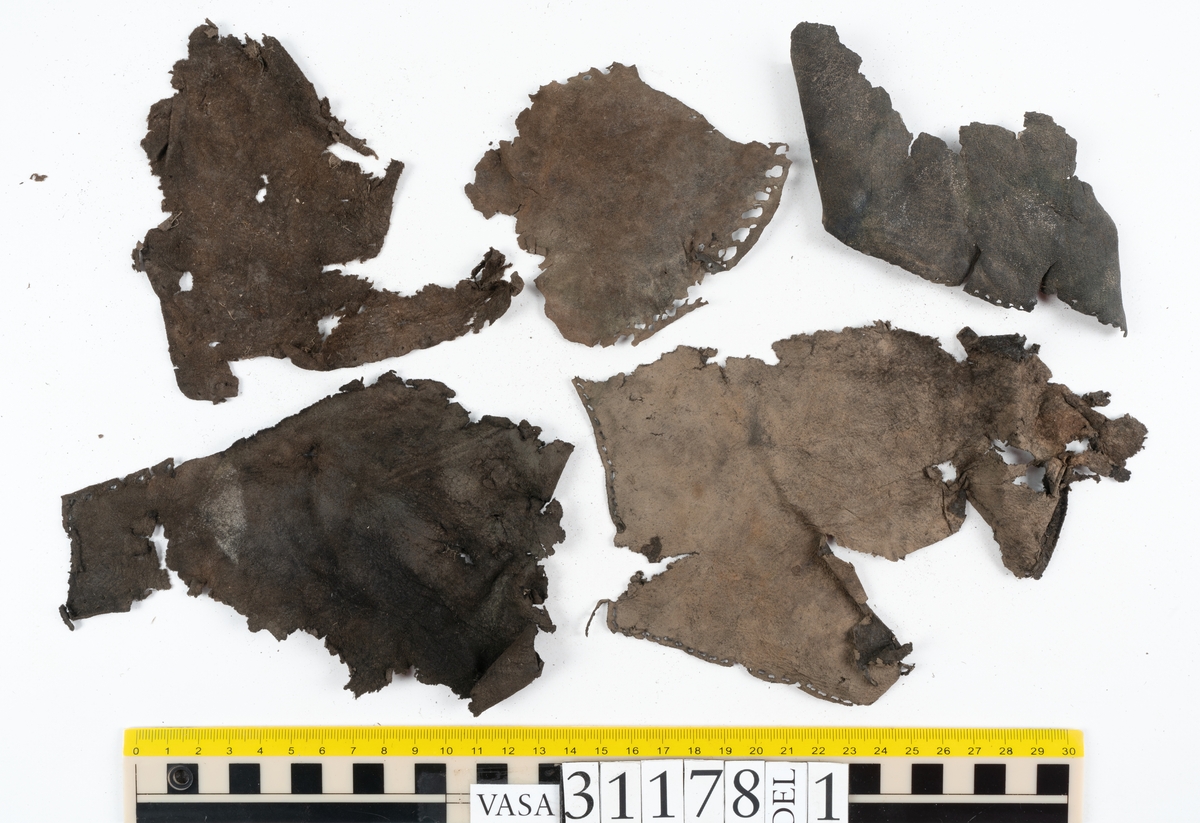 Runt 400 delar och fragment från skor i form av sulor, inlägg, ovanläder, klack, sidostycken, bakkappor, träpliggar, tråd mm. Troligtvis rör det sig om delar och fragment från flera skor och/eller stövlar. Delar av lädret har förstärkts med lim av konserveringsenheten. Merparten av lädret är spaltat samt har hål efter träpligg eller stygn.