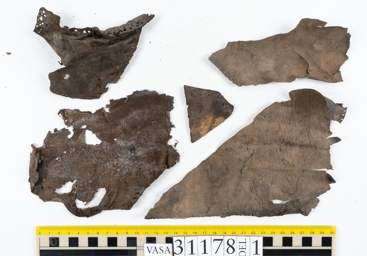 Runt 400 delar och fragment från skor i form av sulor, inlägg, ovanläder, klack, sidostycken, bakkappor, träpliggar, tråd mm. Troligtvis rör det sig om delar och fragment från flera skor och/eller stövlar. Delar av lädret har förstärkts med lim av konserveringsenheten. Merparten av lädret är spaltat samt har hål efter träpligg eller stygn.