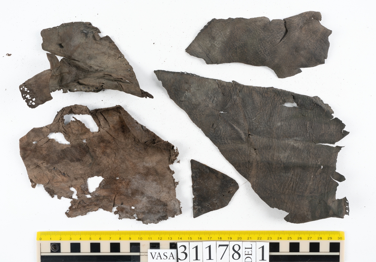 Runt 400 delar och fragment från skor i form av sulor, inlägg, ovanläder, klack, sidostycken, bakkappor, träpliggar, tråd mm. Troligtvis rör det sig om delar och fragment från flera skor och/eller stövlar. Delar av lädret har förstärkts med lim av konserveringsenheten. Merparten av lädret är spaltat samt har hål efter träpligg eller stygn.