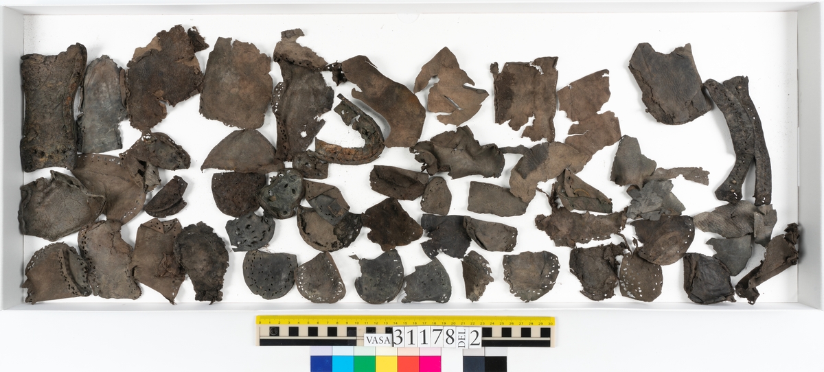Runt 400 delar och fragment från skor i form av sulor, inlägg, ovanläder, klack, sidostycken, bakkappor, träpliggar, tråd mm. Troligtvis rör det sig om delar och fragment från flera skor och/eller stövlar. Delar av lädret har förstärkts med lim av konserveringsenheten. Merparten av lädret är spaltat samt har hål efter träpligg eller stygn.