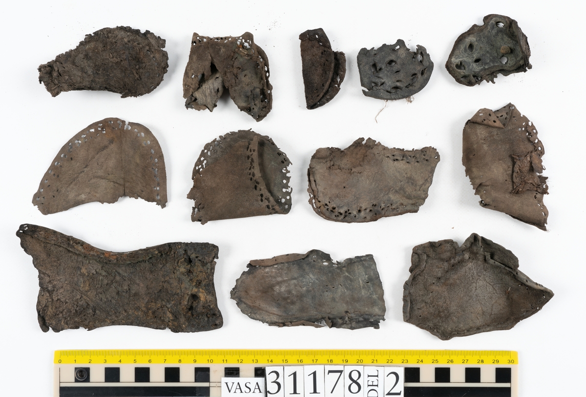 Runt 400 delar och fragment från skor i form av sulor, inlägg, ovanläder, klack, sidostycken, bakkappor, träpliggar, tråd mm. Troligtvis rör det sig om delar och fragment från flera skor och/eller stövlar. Delar av lädret har förstärkts med lim av konserveringsenheten. Merparten av lädret är spaltat samt har hål efter träpligg eller stygn.