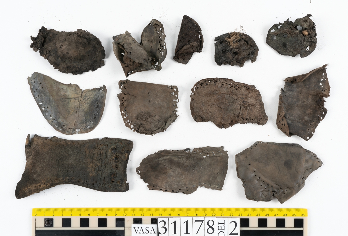 Runt 400 delar och fragment från skor i form av sulor, inlägg, ovanläder, klack, sidostycken, bakkappor, träpliggar, tråd mm. Troligtvis rör det sig om delar och fragment från flera skor och/eller stövlar. Delar av lädret har förstärkts med lim av konserveringsenheten. Merparten av lädret är spaltat samt har hål efter träpligg eller stygn.