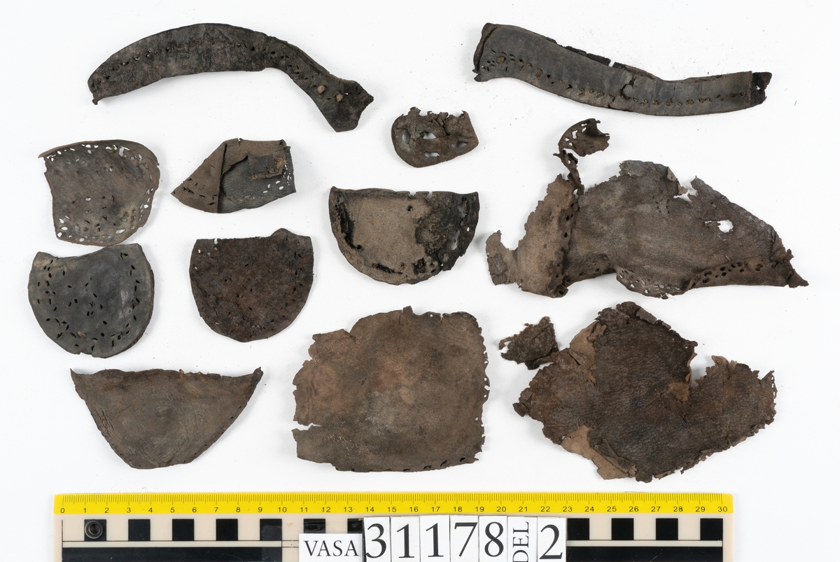 Runt 400 delar och fragment från skor i form av sulor, inlägg, ovanläder, klack, sidostycken, bakkappor, träpliggar, tråd mm. Troligtvis rör det sig om delar och fragment från flera skor och/eller stövlar. Delar av lädret har förstärkts med lim av konserveringsenheten. Merparten av lädret är spaltat samt har hål efter träpligg eller stygn.
