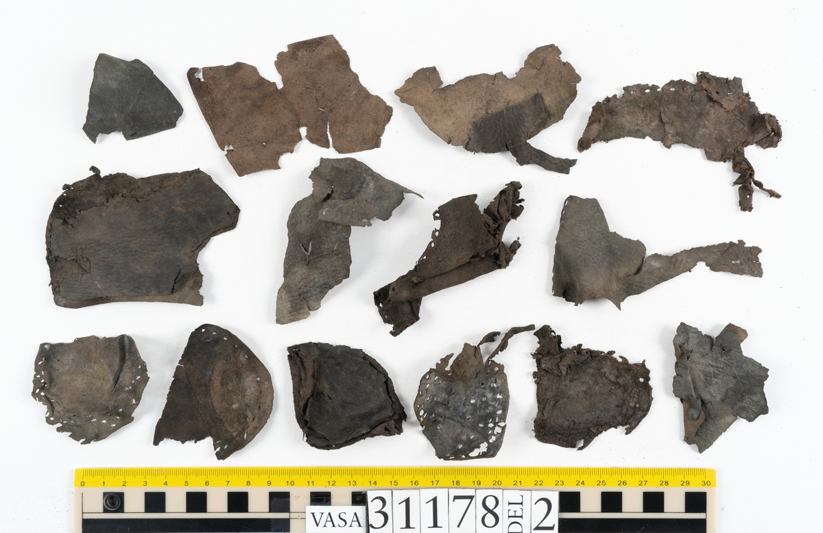 Runt 400 delar och fragment från skor i form av sulor, inlägg, ovanläder, klack, sidostycken, bakkappor, träpliggar, tråd mm. Troligtvis rör det sig om delar och fragment från flera skor och/eller stövlar. Delar av lädret har förstärkts med lim av konserveringsenheten. Merparten av lädret är spaltat samt har hål efter träpligg eller stygn.