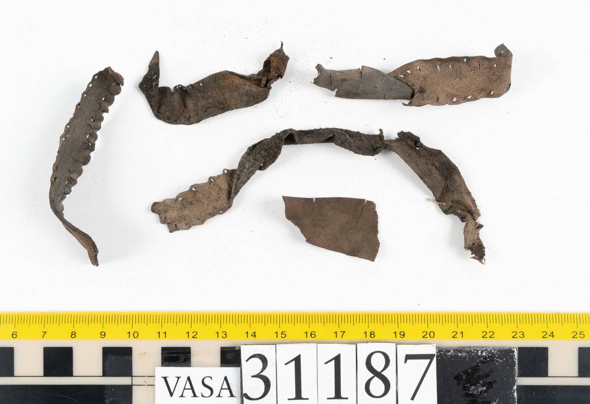 En stropp samt delar av stroppar till ett par stövlar. Den bäst bevarade stroppen är vikt dubbel. Ändarna är spetsiga samt har hål efter stygn. Totalt rör det sig om delar till minst tre stroppar med spetsiga ändar. Lädret ligger vikt samt är sprött och fragmentariskt.