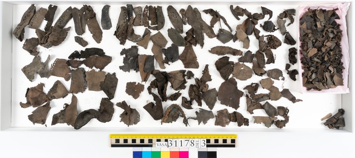 Runt 400 delar och fragment från skor i form av sulor, inlägg, ovanläder, klack, sidostycken, bakkappor, träpliggar, tråd mm. Troligtvis rör det sig om delar och fragment från flera skor och/eller stövlar. Delar av lädret har förstärkts med lim av konserveringsenheten. Merparten av lädret är spaltat samt har hål efter träpligg eller stygn.