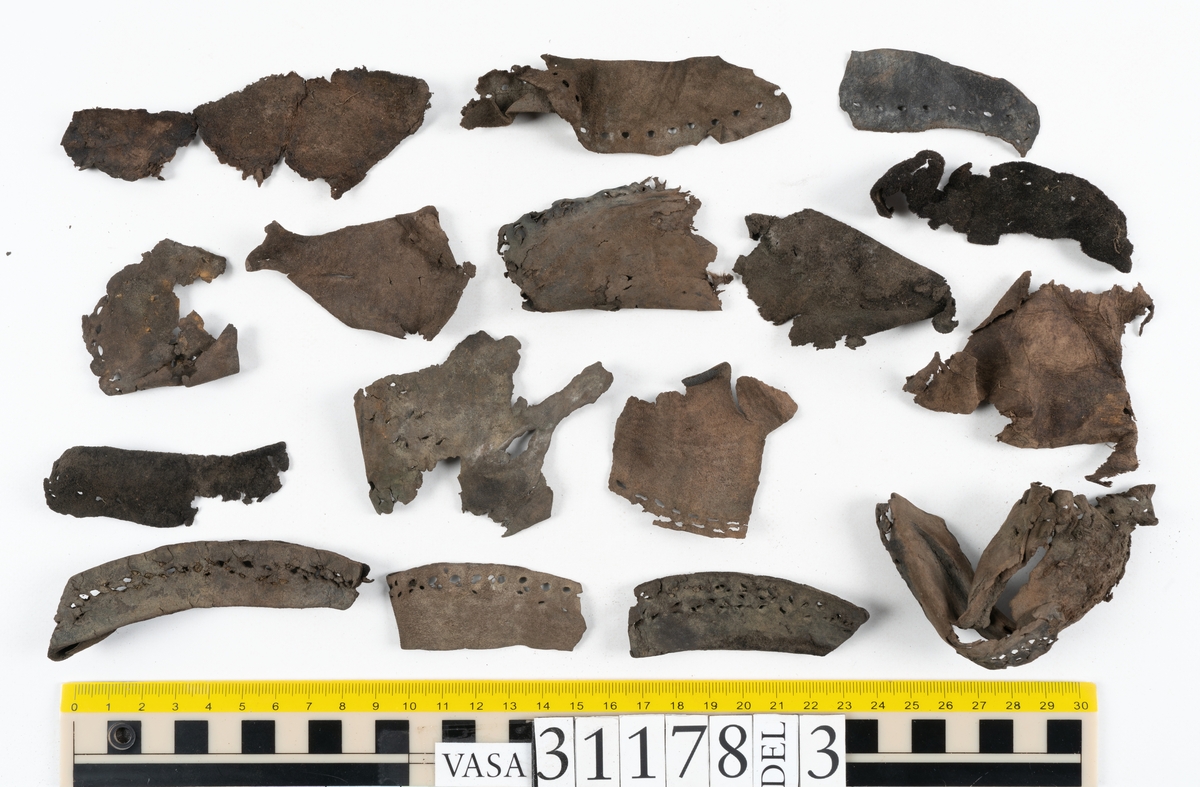 Runt 400 delar och fragment från skor i form av sulor, inlägg, ovanläder, klack, sidostycken, bakkappor, träpliggar, tråd mm. Troligtvis rör det sig om delar och fragment från flera skor och/eller stövlar. Delar av lädret har förstärkts med lim av konserveringsenheten. Merparten av lädret är spaltat samt har hål efter träpligg eller stygn.