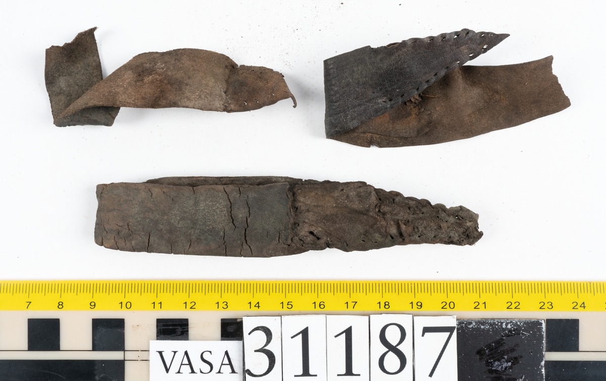 En stropp samt delar av stroppar till ett par stövlar. Den bäst bevarade stroppen är vikt dubbel. Ändarna är spetsiga samt har hål efter stygn. Totalt rör det sig om delar till minst tre stroppar med spetsiga ändar. Lädret ligger vikt samt är sprött och fragmentariskt.