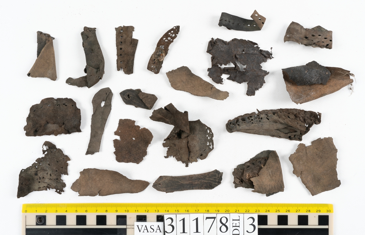 Runt 400 delar och fragment från skor i form av sulor, inlägg, ovanläder, klack, sidostycken, bakkappor, träpliggar, tråd mm. Troligtvis rör det sig om delar och fragment från flera skor och/eller stövlar. Delar av lädret har förstärkts med lim av konserveringsenheten. Merparten av lädret är spaltat samt har hål efter träpligg eller stygn.