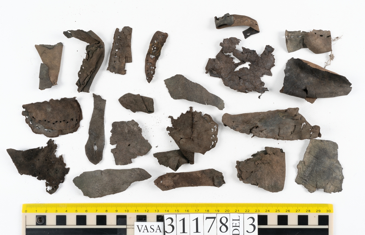 Runt 400 delar och fragment från skor i form av sulor, inlägg, ovanläder, klack, sidostycken, bakkappor, träpliggar, tråd mm. Troligtvis rör det sig om delar och fragment från flera skor och/eller stövlar. Delar av lädret har förstärkts med lim av konserveringsenheten. Merparten av lädret är spaltat samt har hål efter träpligg eller stygn.
