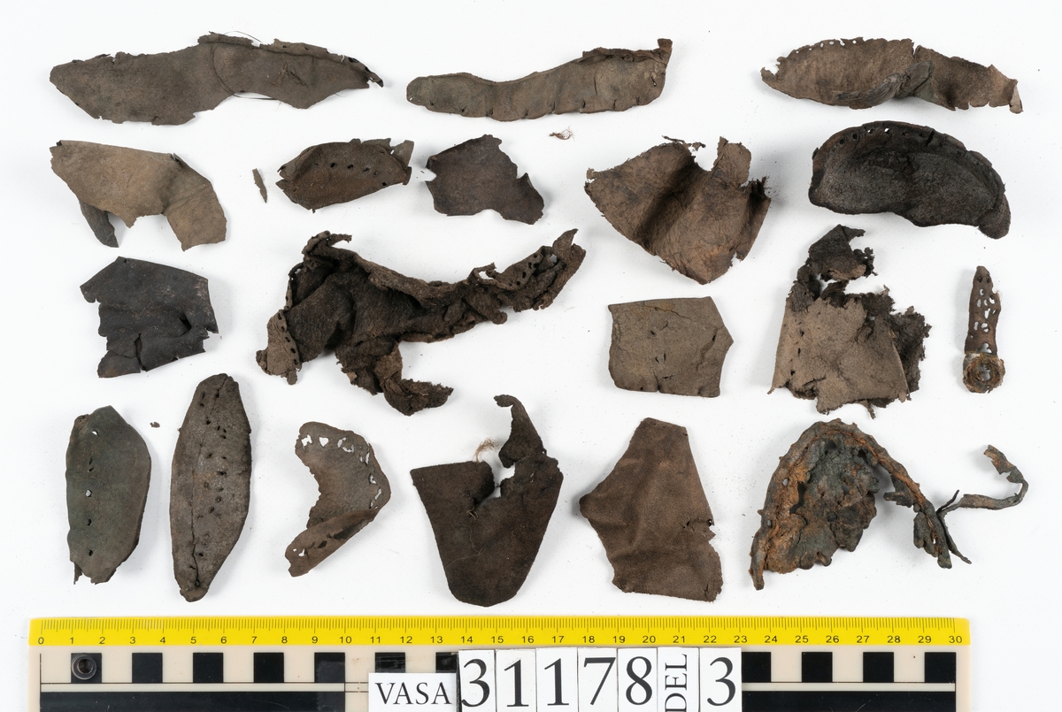 Runt 400 delar och fragment från skor i form av sulor, inlägg, ovanläder, klack, sidostycken, bakkappor, träpliggar, tråd mm. Troligtvis rör det sig om delar och fragment från flera skor och/eller stövlar. Delar av lädret har förstärkts med lim av konserveringsenheten. Merparten av lädret är spaltat samt har hål efter träpligg eller stygn.