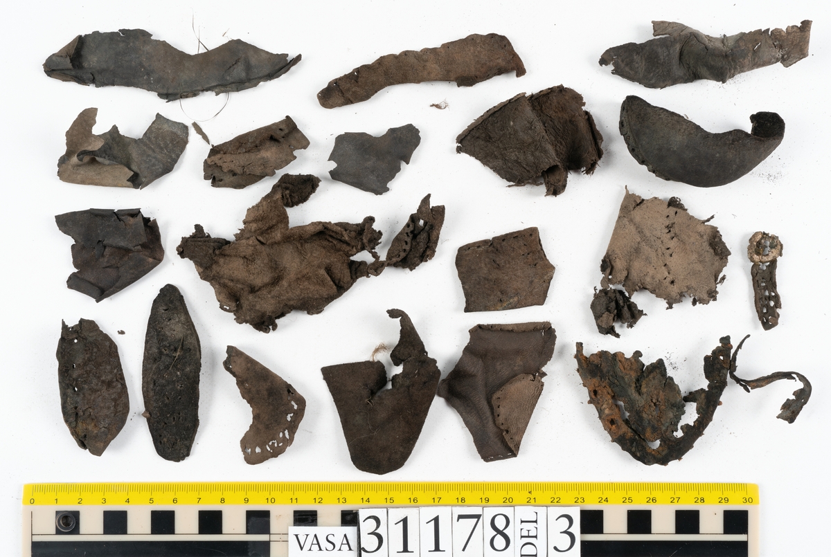 Runt 400 delar och fragment från skor i form av sulor, inlägg, ovanläder, klack, sidostycken, bakkappor, träpliggar, tråd mm. Troligtvis rör det sig om delar och fragment från flera skor och/eller stövlar. Delar av lädret har förstärkts med lim av konserveringsenheten. Merparten av lädret är spaltat samt har hål efter träpligg eller stygn.