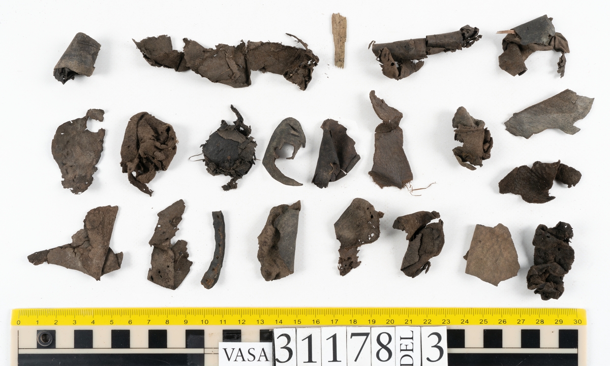 Runt 400 delar och fragment från skor i form av sulor, inlägg, ovanläder, klack, sidostycken, bakkappor, träpliggar, tråd mm. Troligtvis rör det sig om delar och fragment från flera skor och/eller stövlar. Delar av lädret har förstärkts med lim av konserveringsenheten. Merparten av lädret är spaltat samt har hål efter träpligg eller stygn.