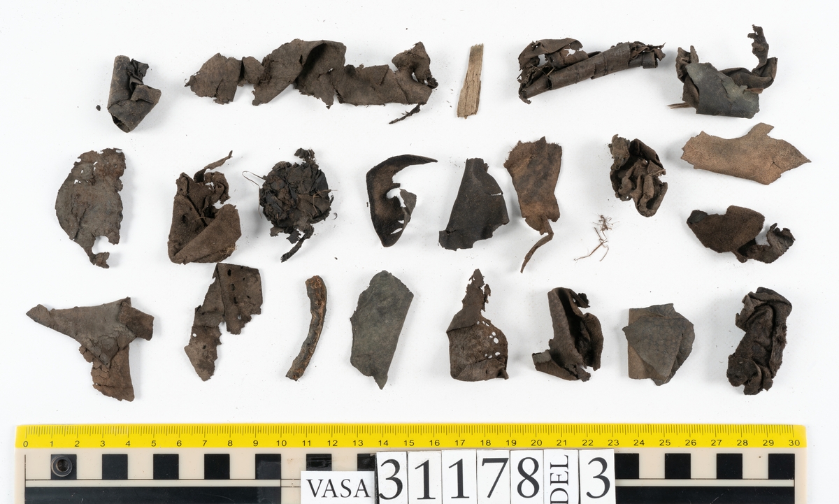 Runt 400 delar och fragment från skor i form av sulor, inlägg, ovanläder, klack, sidostycken, bakkappor, träpliggar, tråd mm. Troligtvis rör det sig om delar och fragment från flera skor och/eller stövlar. Delar av lädret har förstärkts med lim av konserveringsenheten. Merparten av lädret är spaltat samt har hål efter träpligg eller stygn.