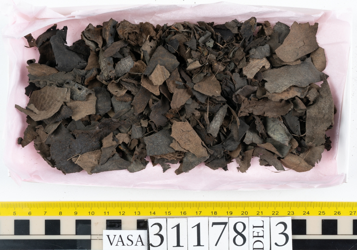 Runt 400 delar och fragment från skor i form av sulor, inlägg, ovanläder, klack, sidostycken, bakkappor, träpliggar, tråd mm. Troligtvis rör det sig om delar och fragment från flera skor och/eller stövlar. Delar av lädret har förstärkts med lim av konserveringsenheten. Merparten av lädret är spaltat samt har hål efter träpligg eller stygn.