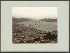 Bilden föreställer en panorama från Nagasaki. Den är tagen från en förhöjd ståndpunkt på kullarna ovan på staden. I förgrunden syns staden låga träbebyggelse och i bakgrunden bukten och hamnen fullt med båtar och skepp.