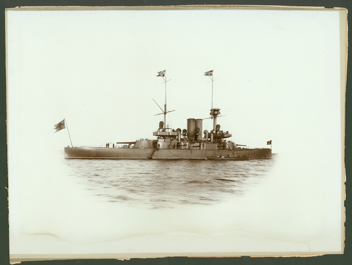 Fartygsporträtt som visar pansarbåten Oden till ankars. Fartyget syns från babordssidan där ett mindre ångbåt ligger förtöjt. Flera repstege leder upp till däcket. På akterdäcket står tre men som verka samtala.