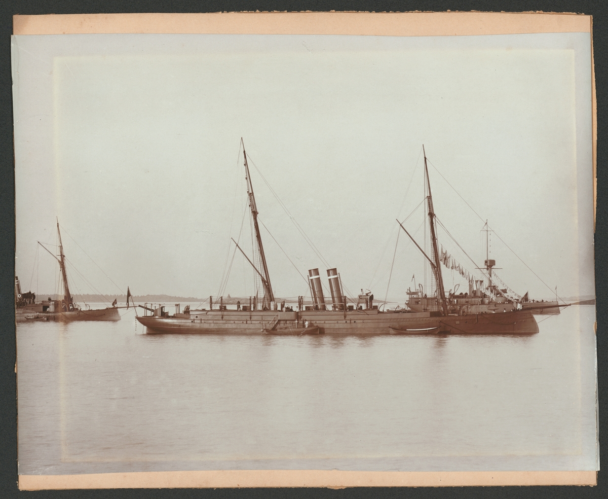 Fartygsporträtt som visar kanonbåten Edda till ankars. Fartyget syns från babordssidan där två skeppsbåtar ligger förtöjt. I bakgrunden syns ett skärgårdslandskap och ytterligare två kanonbåtar.