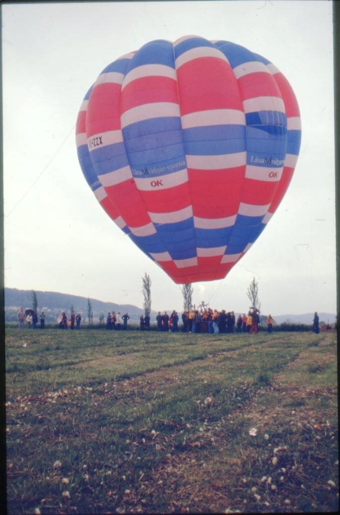 En röd-vit-blå ballong märkt "OK" står färdig att lyfta, brännaren är igång. En folksamling runt ballongkorgen.