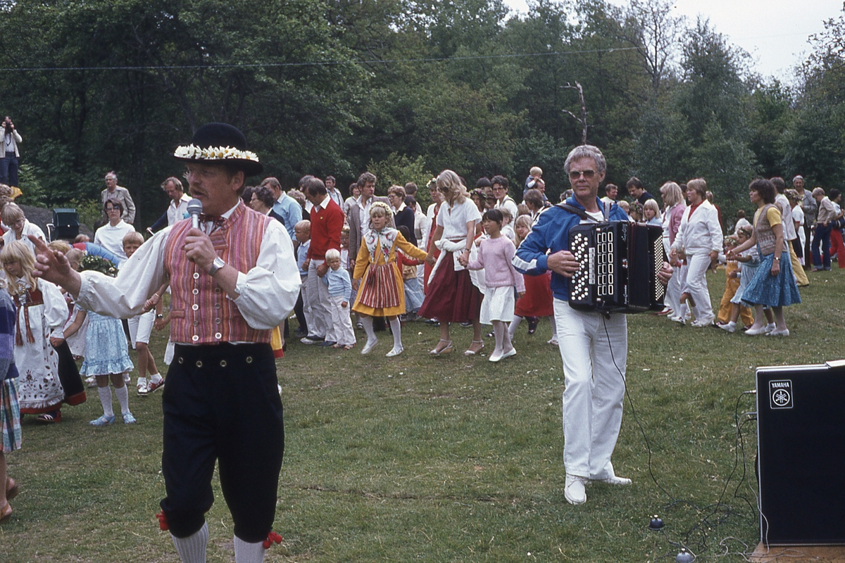 Barn och vuxna dansar kring stången vid midsommarfirande på Ekensås cirka 1982. Sångare/ledare är Olle Svensson (klädd i folkdräkt) samt spelmannen Frank Efraimsson som spelar dragspel.