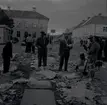 Torget på kvällen efter marknaden i Mörbylånga. Män i kostym samt en kvinna och barn som står i skräp på marken. Åbrinks i bakgrunden.