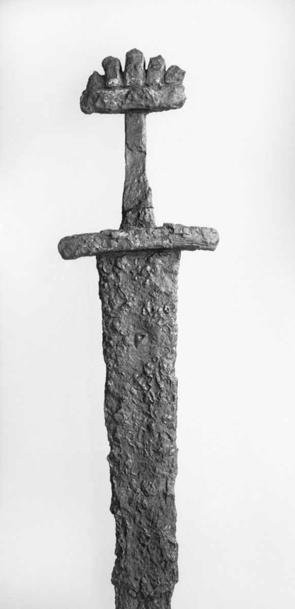 Tveegget sverd med femtunget knap, type RYGH 507. Hjalt og knap av jern, men spor av sølv og ornamenter. Klingen adskillig skadet og det hele stygt forrustet. Længden nu 77 cm., og et avbrutt stykke av klingen 16,8 cm. langt
Revisjon 2019: Tveegget sverd som Petersens type O. Odden mangler, og nederste del av klingen er brukket av, men er tilstede. Klingen er bøyd og vridd i nederste halvdel, her er det også store hakk. Underhjaltet er svakt nedoverbøys, og har spor til innlagte metalltråder. På begge hjalt samt knappen har det vært innlagt dekor i form av et flettemønster, og dekoren er delvis bevart, men er utydelig. Trolig er sverdet av Petersens undergruppe I. 