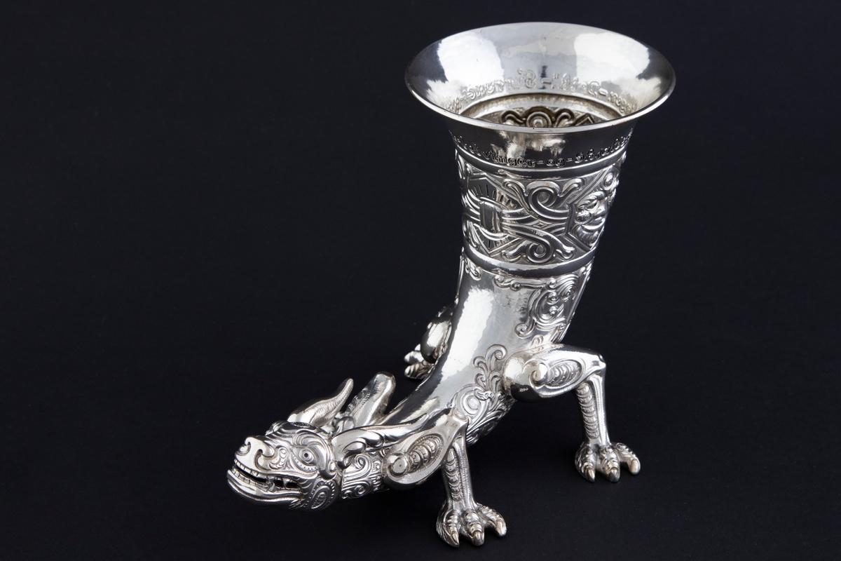 Drikkehorn i sølv som hviler på fire føtter med klør. Enden av hornet er utformet som et dragehode, mens øvre del er dekorert med to middelalderske masker; den ene smiler, mens den andre har et sint uttrykk. Yttersiden er dekorert med flettverksornamentikk, som trolig opprinnelig har vært oksidert for å forsterke kontrasten til de blanke partiene. Innsiden har rester av forgylling.