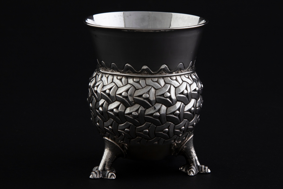 Sølvbeger. Kuleformet korpus hvis ytterside er dekorert med "entrelacs" (sammenkjedete trekanter med konkave sider). Det bæres av tre støpte drageben. Øvre del av begeret er konisk med bølgeformet dekor nederst.