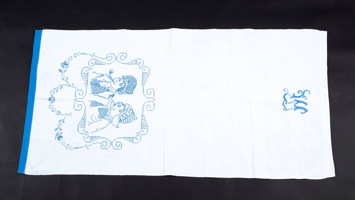 Pyntehåndkle, Hvit bunn, blå kanter. Brodert med en gutt og ei jente med korsting. Monogram M.

Gitt av Erna Madsen. Produsert av Margit Madsen.
Disse drev manufaktur i Drøbak.
