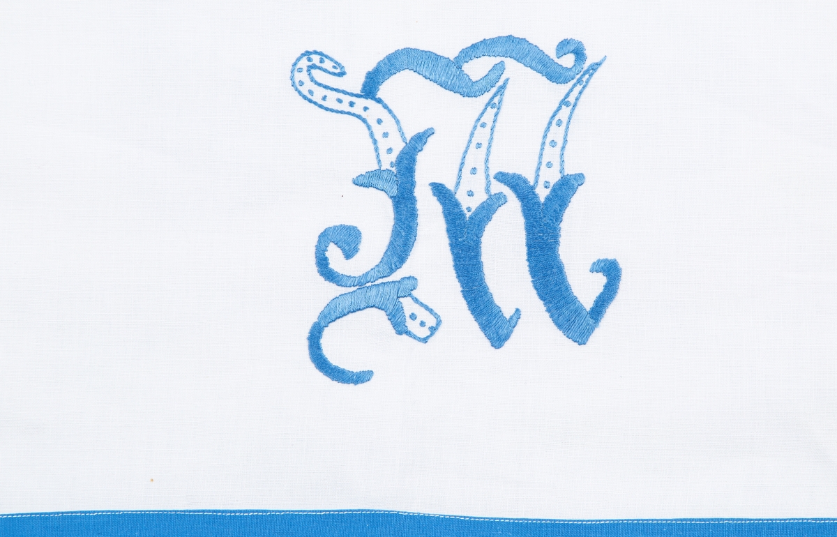 Pyntehåndkle, Hvit bunn, blå kanter. Brodert med en gutt og ei jente med korsting. Monogram M.

Gitt av Erna Madsen. Produsert av Margit Madsen.
Disse drev manufaktur i Drøbak.