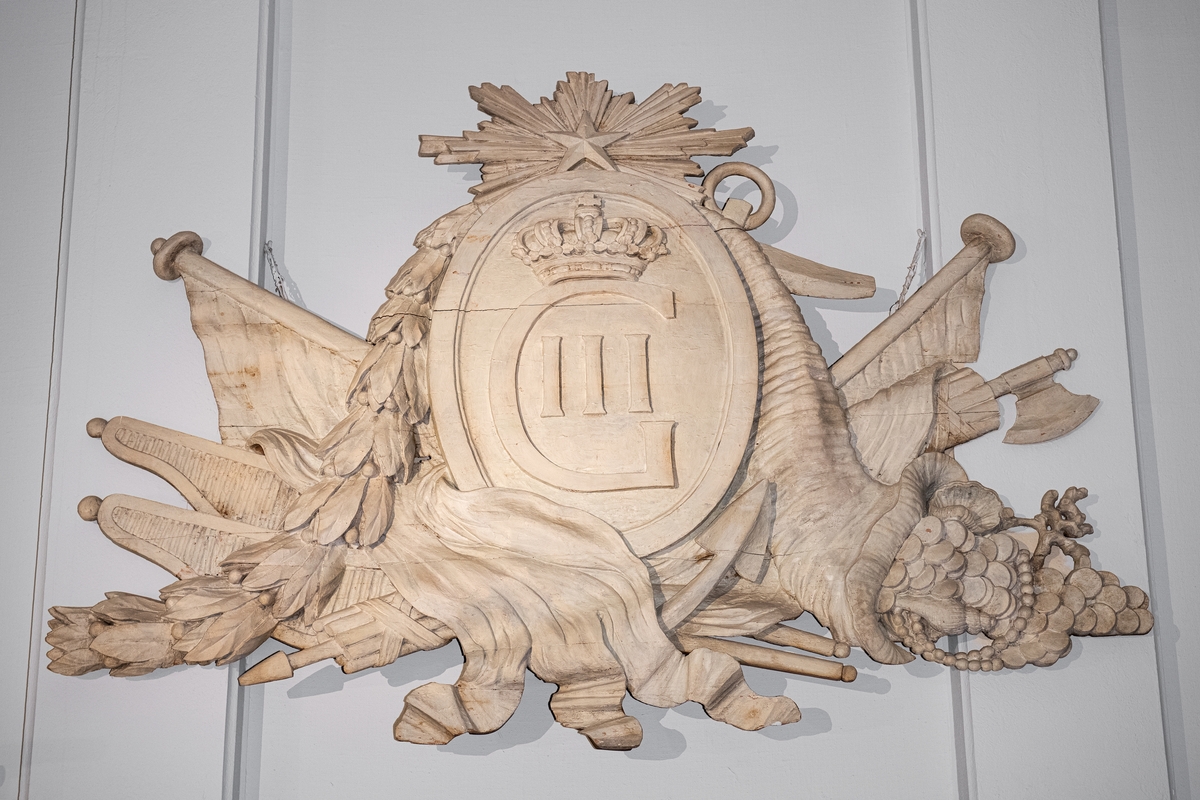 Akterspegelsornament från linjeskeppet Gustaf III. Vapensköld med Gustaf III:s krönta namnschiffer med stjärna och strålknippe över och diverse emblem såsom flaggor, ankare, ymnighetshorn, pilkoger, spöknippe och bladslinga å sidorna.
