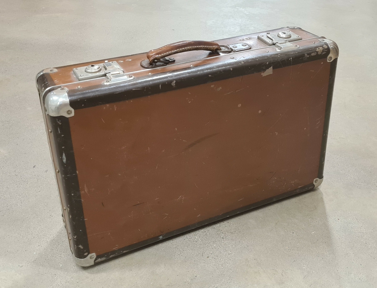 Resväska av brunt unikamaterial. Väskan är försedd med metallförstärkta hörn samt två lås. På väskan sitter en klisterlapp där det står Hotel Moltke Grand Hotell de Rome, Berlin.