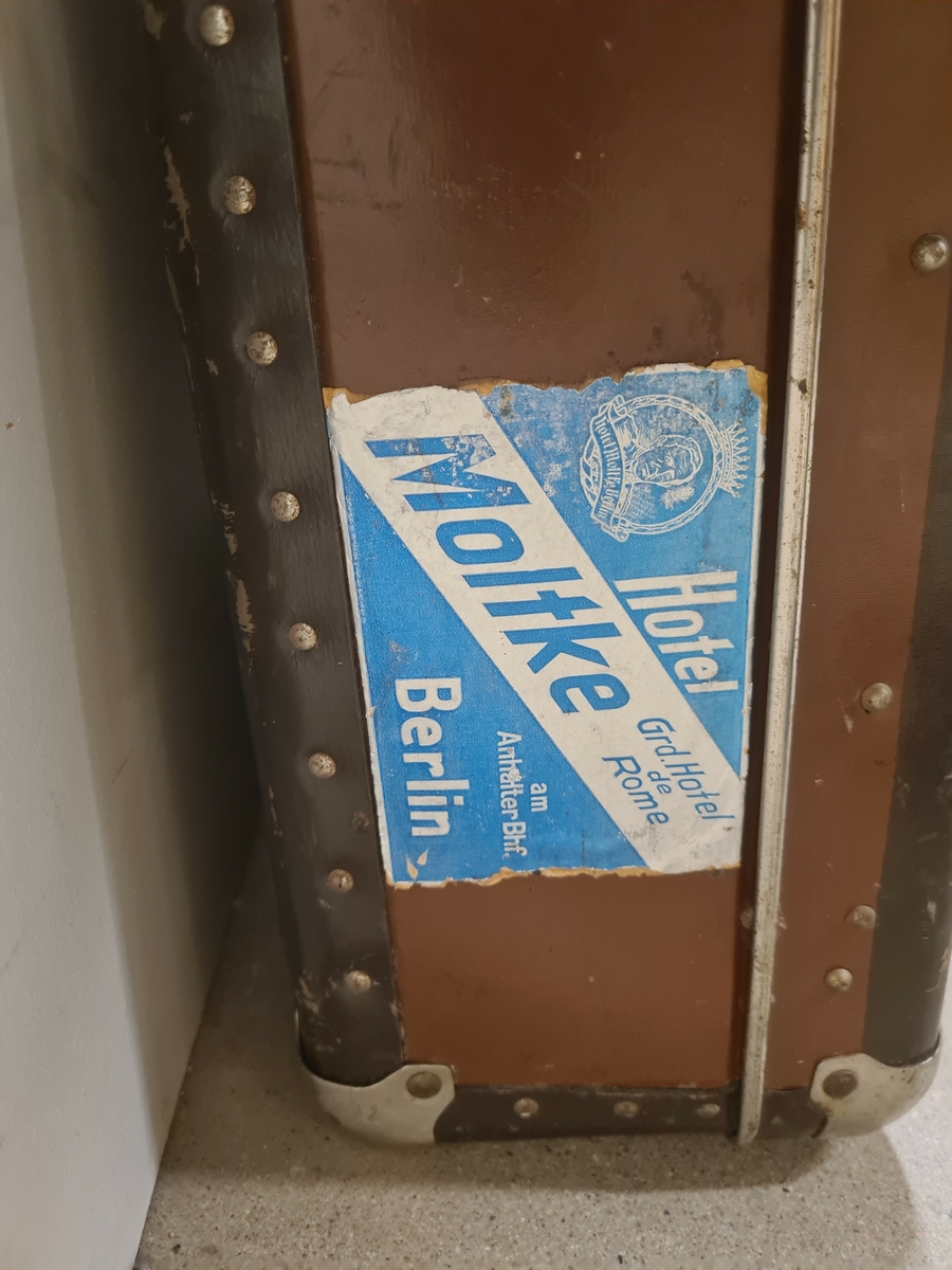 Resväska av brunt unikamaterial. Väskan är försedd med metallförstärkta hörn samt två lås. På väskan sitter en klisterlapp där det står Hotel Moltke Grand Hotell de Rome, Berlin.